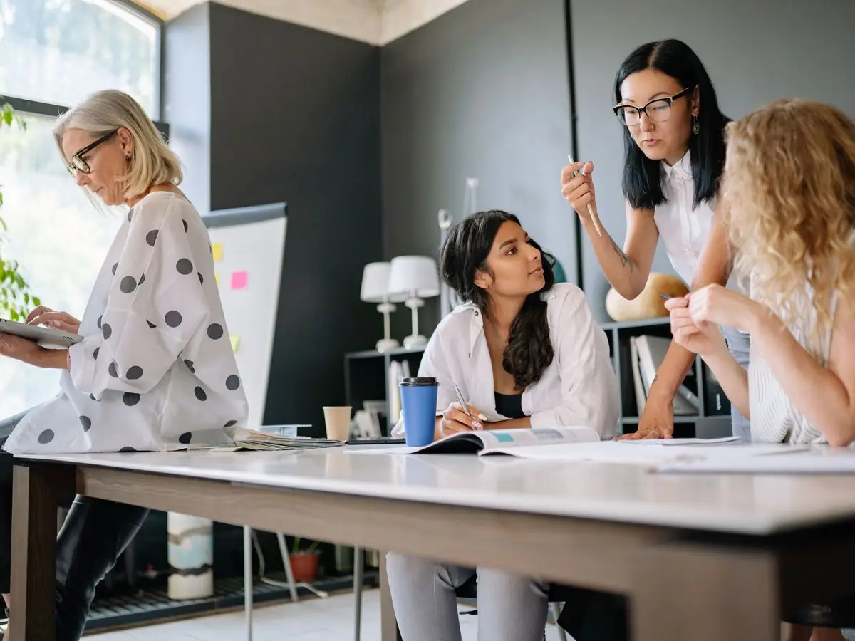 Four colleagues work together at an office table, one on a tablet, two looking over a document with the person writing.