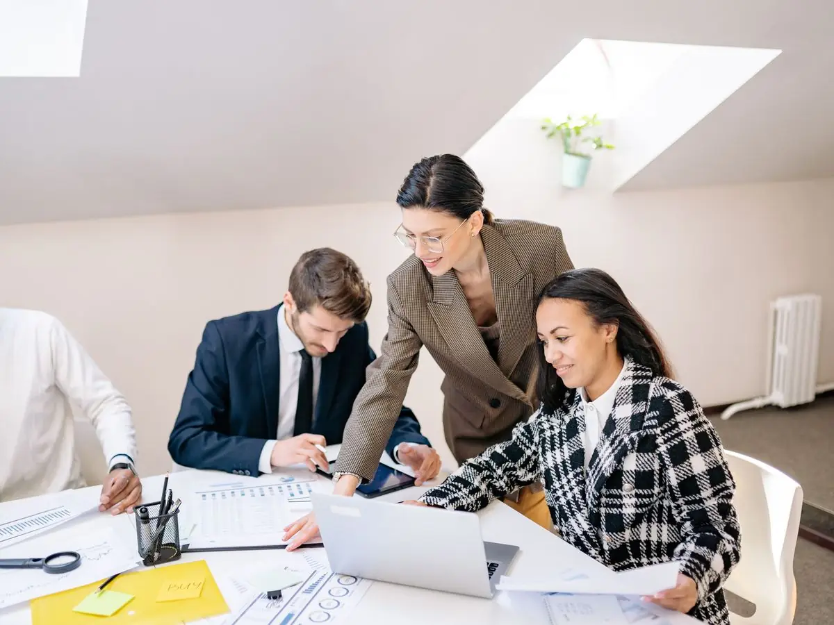 A team lead leans over an office table to go through the work with three colleagues, one of them at a laptop.